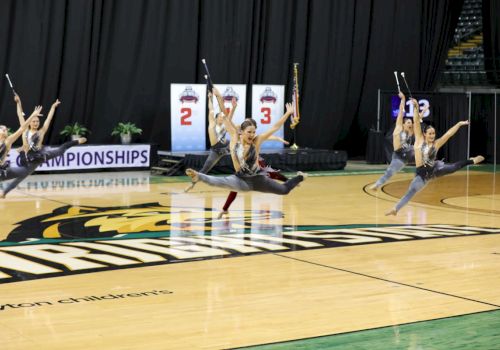 Performers in synchronized dance or baton routine leap in unison on a gym floor.