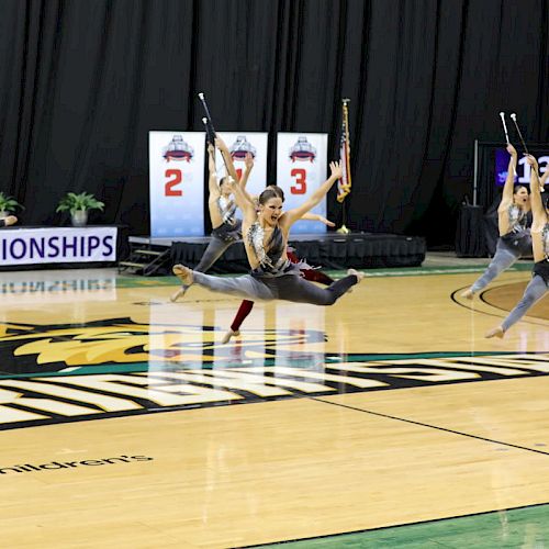 Performers in synchronized dance or baton routine leap in unison on a gym floor.