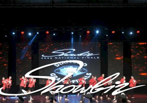 A group of performers in red outfits dance on stage during the "Showbiz 2023 National Finals" with a colorful backdrop and lights.
