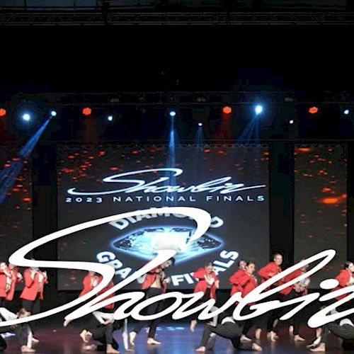 A group of performers in red outfits dance on stage during the "Showbiz 2023 National Finals" with a colorful backdrop and lights.