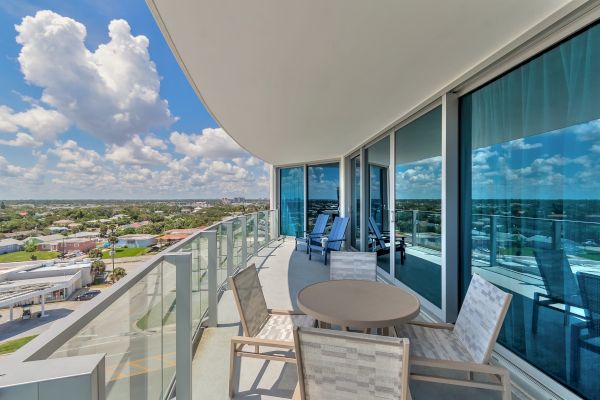 A modern balcony with glass railings, an outdoor dining set, lounge chairs, and a view of a suburban area under a partly cloudy sky can be seen.