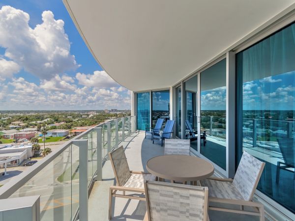 A modern balcony with glass railings, an outdoor dining set, lounge chairs, and a view of a suburban area under a partly cloudy sky can be seen.