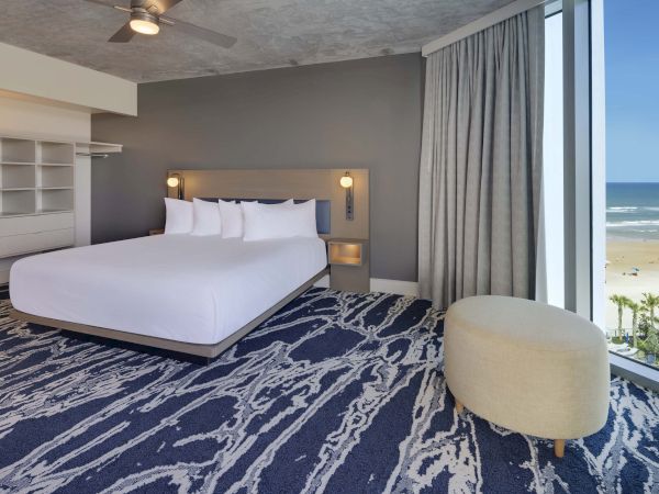 A modern hotel room features a large bed, wall-mounted lamps, a lounge chair, and a scenic ocean view through floor-to-ceiling windows.