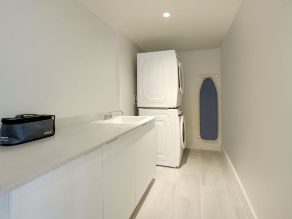 A laundry room with a stacked washer and dryer, a sink, an ironing board on the wall, and a counter with a black storage bag on top.