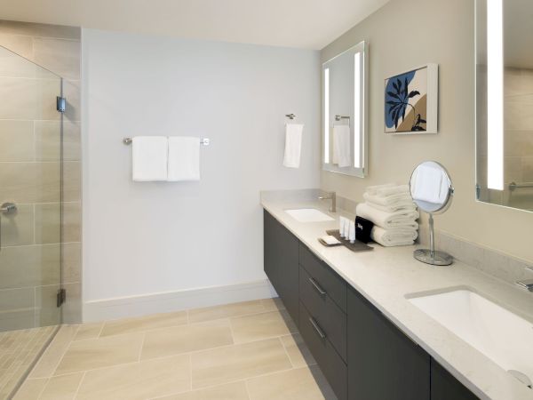 A modern bathroom features a double sink vanity, a walk-in glass shower, a stack of folded towels, artwork, and a small round mirror on the countertop.