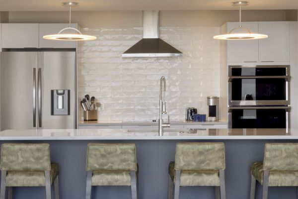 A modern kitchen features a stainless steel refrigerator, oven, and range hood, with a central island and padded stools underneath.