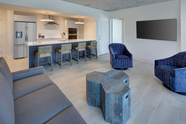 A modern living area with blue chairs, a grey sofa, and unique wooden tables. The kitchen in the background has bar stools and contemporary decor.