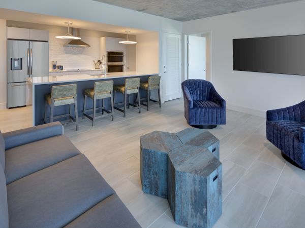 A modern living area with blue chairs, a grey sofa, and unique wooden tables. The kitchen in the background has bar stools and contemporary decor.