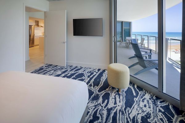 A modern bedroom with a TV, patterned carpet, an ottoman, and sliding doors leading to a balcony with lounge chairs and an ocean view ends the sentence.