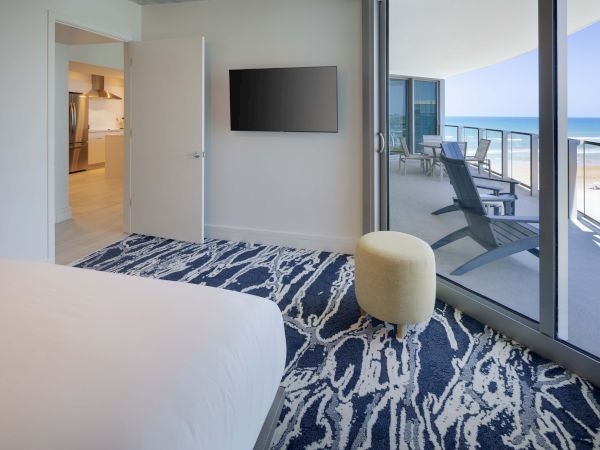 A modern bedroom with a TV, patterned carpet, an ottoman, and sliding doors leading to a balcony with lounge chairs and an ocean view ends the sentence.