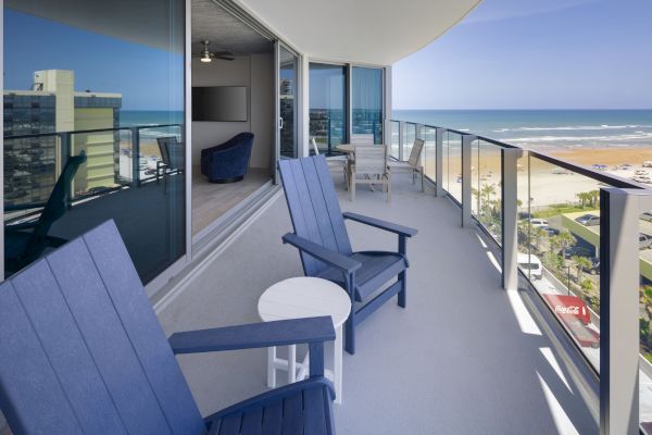 A balcony view with blue chairs, a small white table, glass railing, overlooking the beach and ocean with high-rise buildings nearby, in daytime.