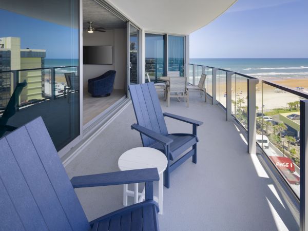 A balcony view with blue chairs, a small white table, glass railing, overlooking the beach and ocean with high-rise buildings nearby, in daytime.
