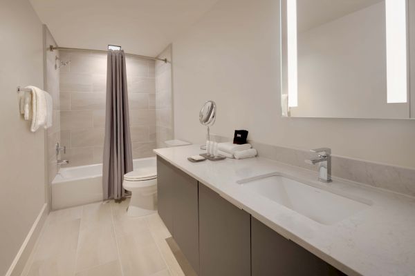 A modern bathroom with a large vanity, a sink, a mirror, towels, a toilet, and a bathtub with a gray shower curtain, featuring sleek design points.