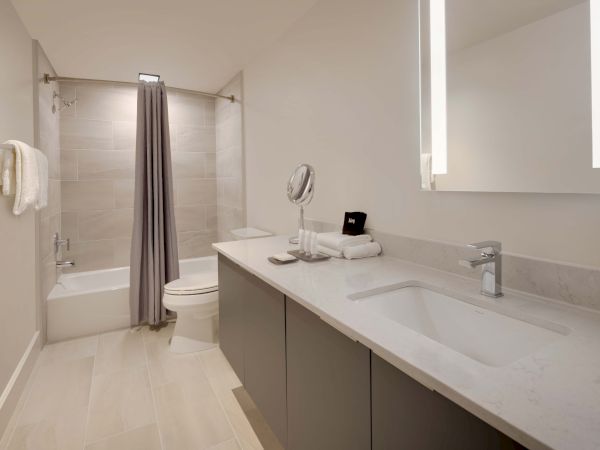 A modern bathroom with a large vanity, a sink, a mirror, towels, a toilet, and a bathtub with a gray shower curtain, featuring sleek design points.