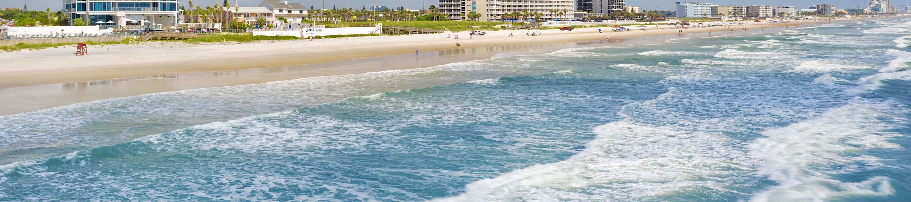 A scenic beach with clear blue waters, waves crashing onto the shore, and several buildings along the coastline on a partly cloudy day.