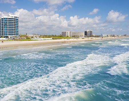 A scenic beach with clear blue waters, waves crashing onto the shore, and several buildings along the coastline on a partly cloudy day.