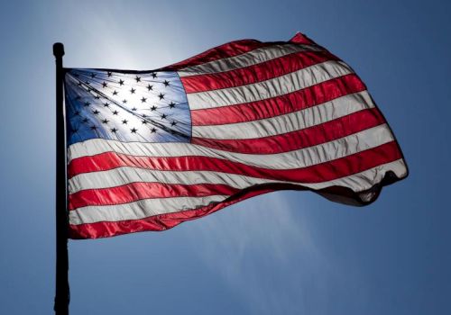The image shows an American flag waving against a blue sky background, with sunlight shining through the fabric.