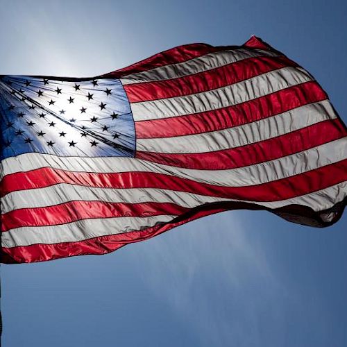 The image shows an American flag waving against a blue sky background, with sunlight shining through the fabric.