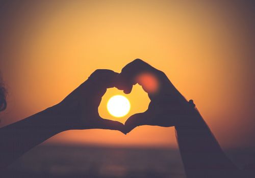 The image shows two hands forming a heart shape with the sun inside the heart, against the backdrop of a sunset or sunrise.