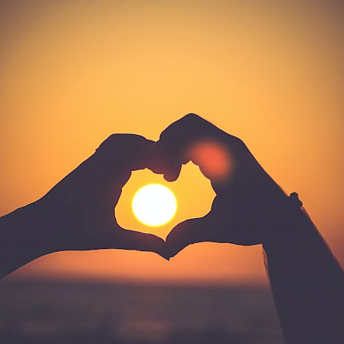 The image shows two hands forming a heart shape with the sun inside the heart, against the backdrop of a sunset or sunrise.