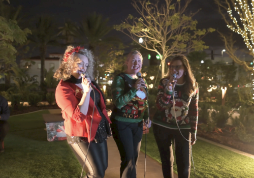Three women sing with microphones outdoors at night, wearing festive sweaters as string lights glow around them.