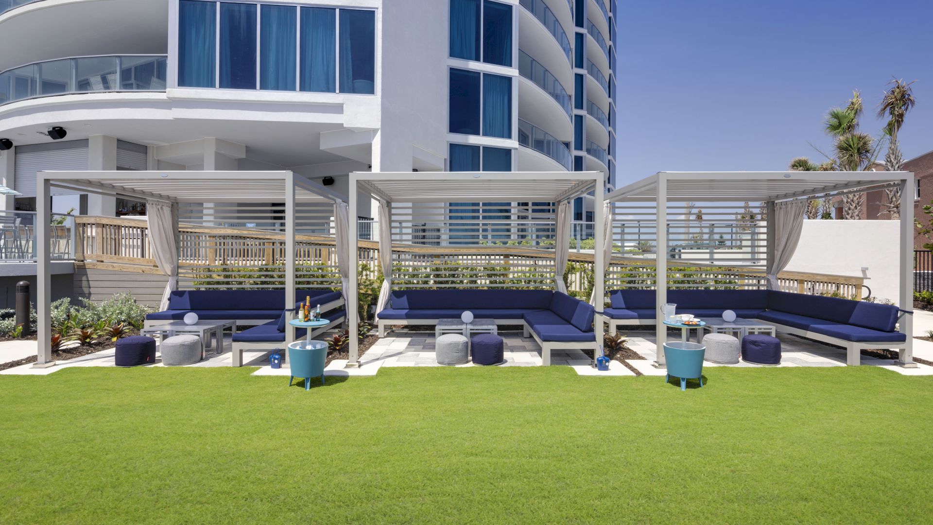 Poolside lounge area with covered seating, blue cushions, small tables, and neatly trimmed grass in front of a modern curved building.