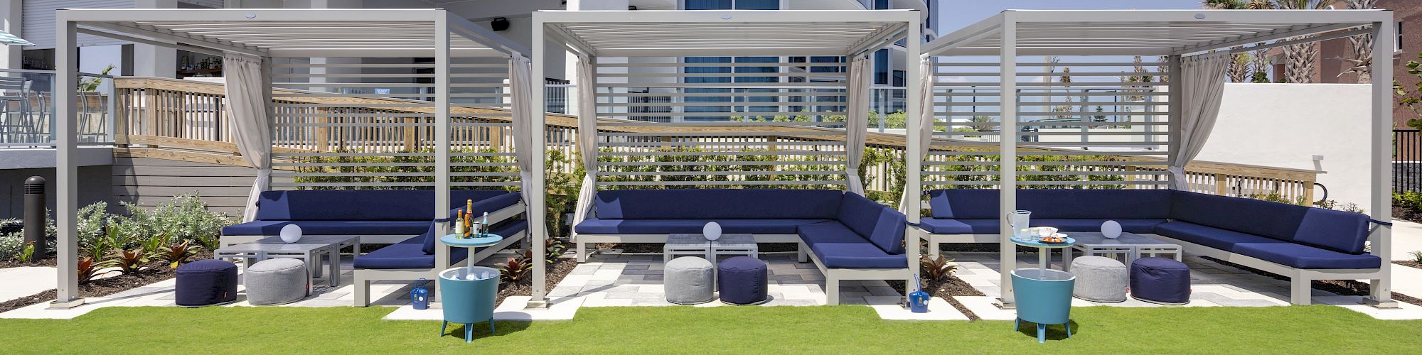 Poolside lounge area with covered seating, blue cushions, small tables, and neatly trimmed grass in front of a modern curved building.