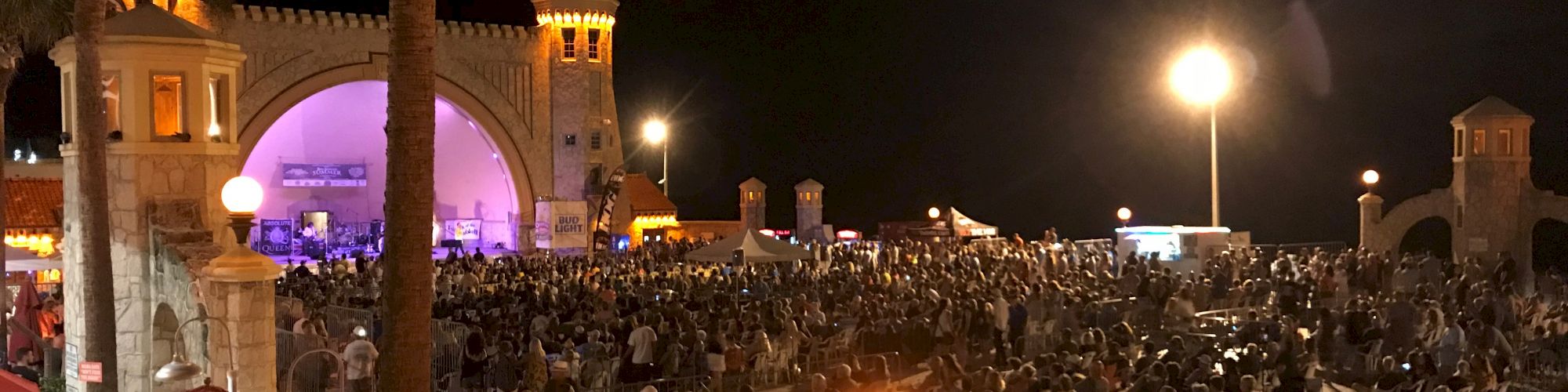 A large crowd gathered at an outdoor event at night, with a stage and lit archway in the background, surrounded by palm trees and streetlights.