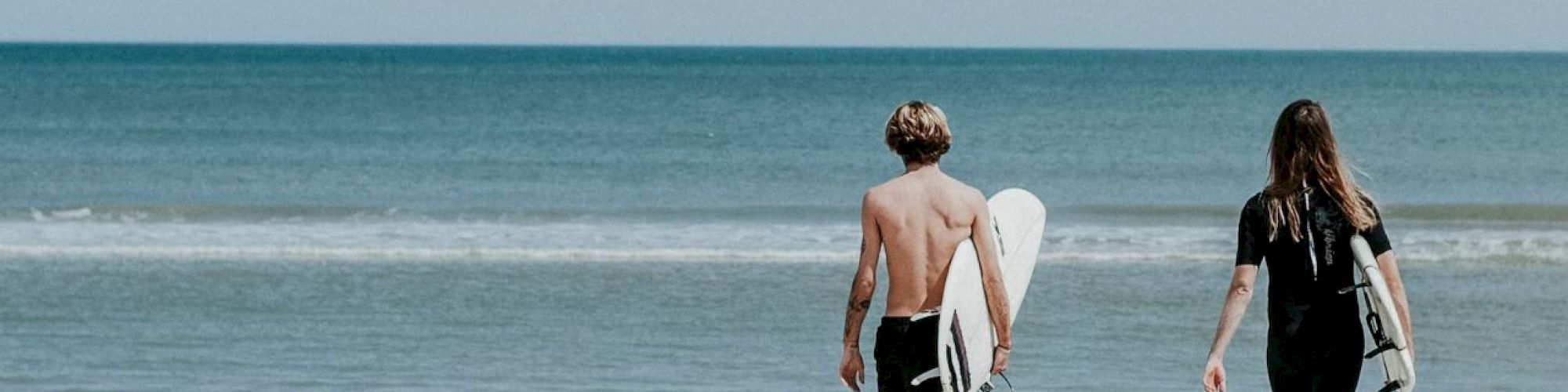 Two people carrying surfboards walk towards the ocean on a sandy beach.