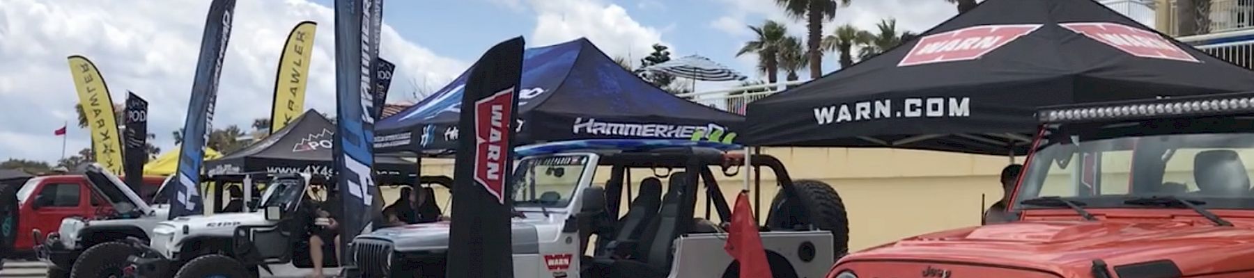 Various customized off-road vehicles are displayed under branded tents and banners at an outdoor event, with palm trees and a partly cloudy sky above.