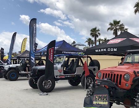 Various customized off-road vehicles are displayed under branded tents and banners at an outdoor event, with palm trees and a partly cloudy sky above.