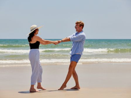 A couple enjoys a sunny day at the beach, holding hands and smiling, with the ocean waves in the background.