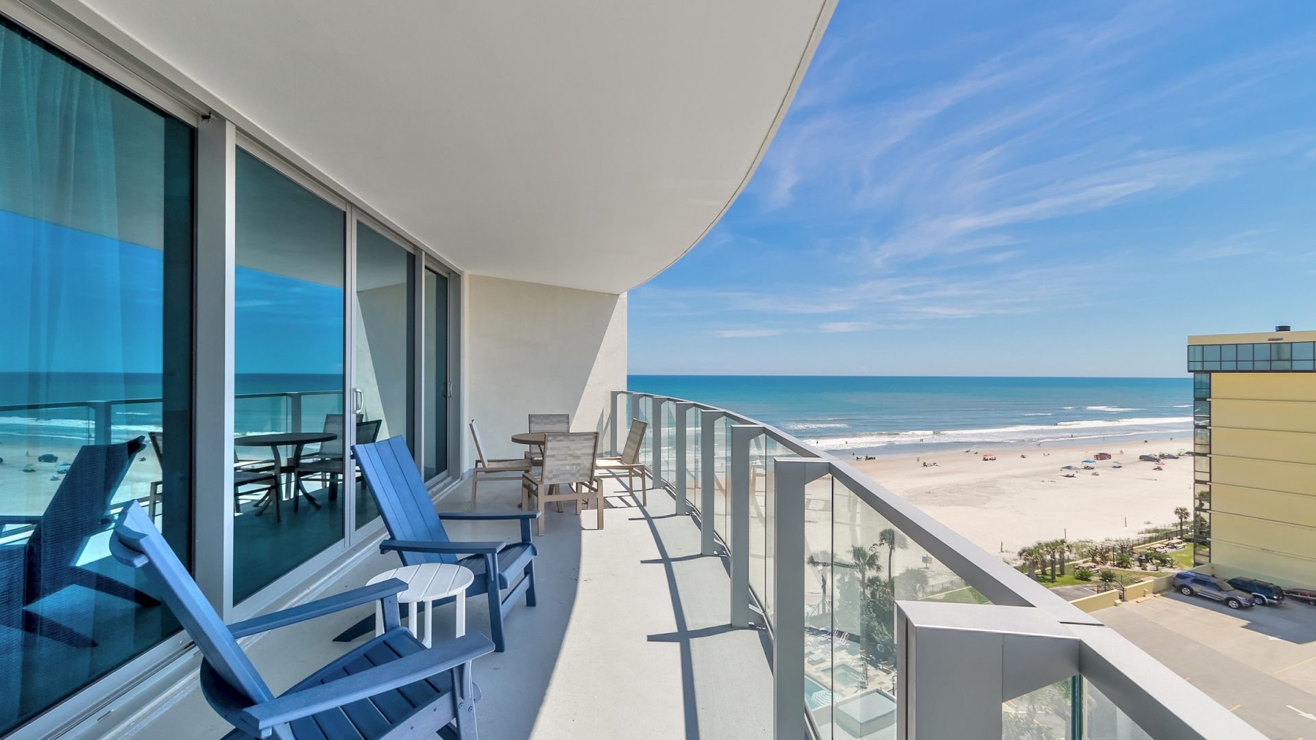 A modern balcony with blue chairs and a table overlooks a sandy beach and distant ocean with a clear sky, adjacent to a neighboring building.
