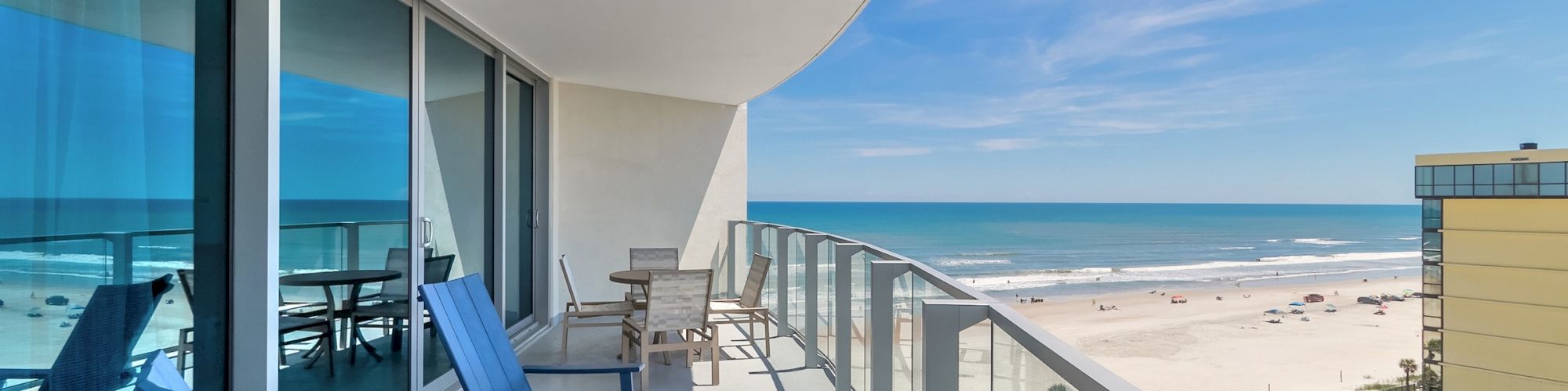 A modern balcony with blue chairs and a table overlooks a sandy beach and distant ocean with a clear sky, adjacent to a neighboring building.