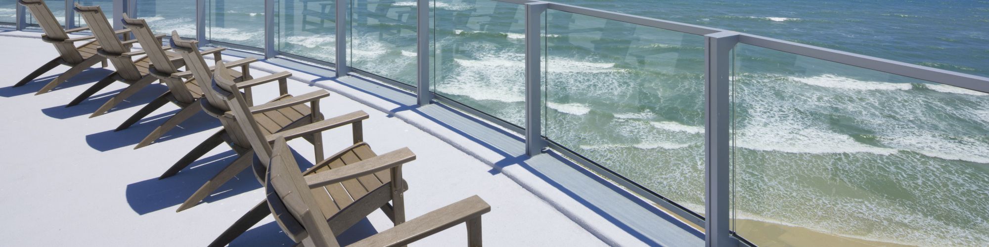 This image shows a row of wooden lounge chairs on a waterfront deck with glass railings overlooking a beach and ocean under a clear, sunny sky.