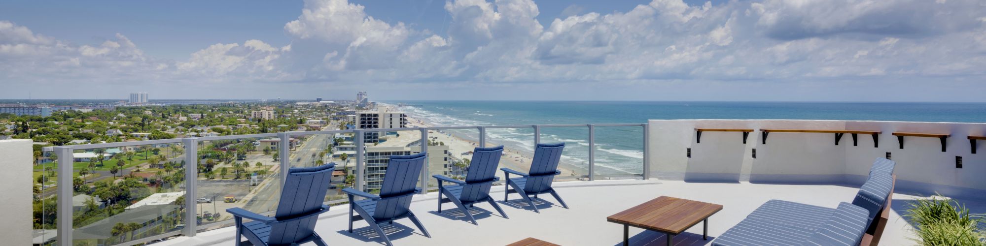 A rooftop terrace with blue lounge chairs and benches overlooking a scenic beachfront and city skyline, under a clear blue sky.