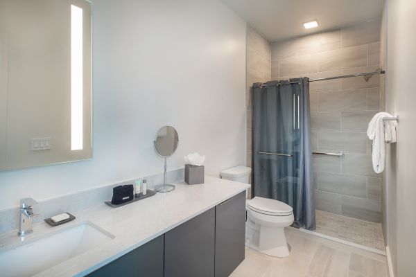 A modern bathroom featuring a sink, mirror, toiletries, toilet, and shower with grey tiles and a curtain, plus neatly arranged towels.