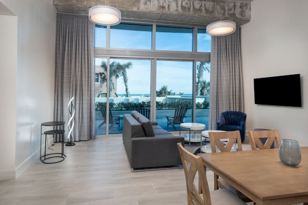 A modern living room with a dining table, couch, TV, and large windows opening to a balcony with a seaside view, palm trees visible.