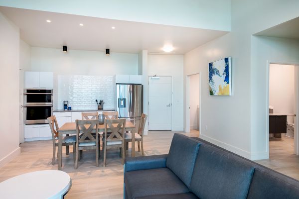 A modern kitchen and living area with a dining table, six chairs, a refrigerator, a stove, a couch, and a colorful painting on the wall.