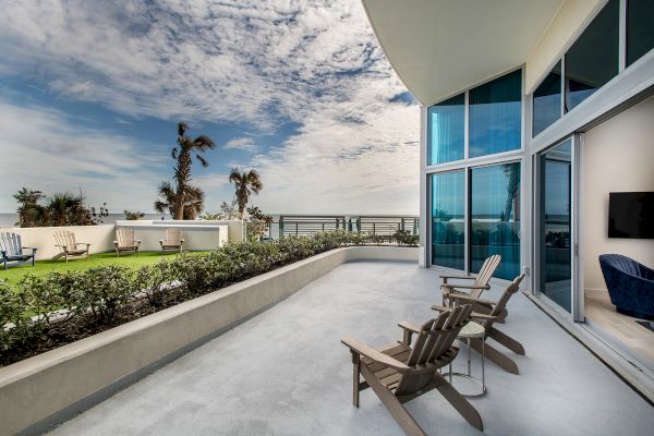 A modern patio with wooden chairs, glass doors, a green garden, and a view of the ocean under a partly cloudy sky is shown in the image.