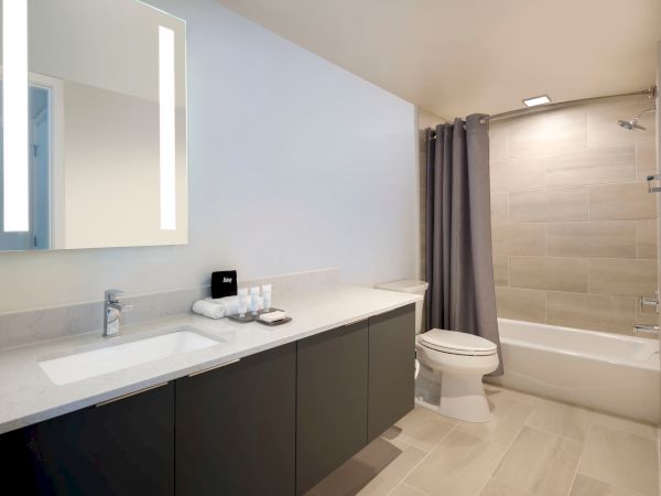 A modern bathroom with a vanity, illuminated mirror, toilet, and a bathtub with a gray shower curtain, tiled walls and floor.