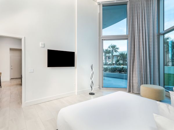 A modern bedroom with a TV on the wall, a bed, large windows with curtains, and a view of an outdoor area with trees and a pool.