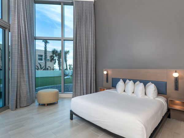 A modern bedroom features a large bed with white linens and multiple pillows, floor-to-ceiling windows with grey curtains, and a small ottoman.