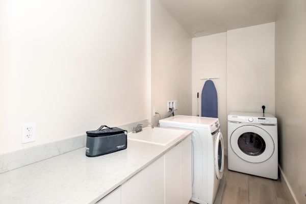 This image shows a laundry room with a washing machine, dryer, countertop, sink, and an ironing board in the corner.