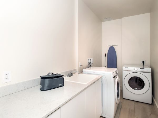 This image shows a laundry room with a washing machine, dryer, countertop, sink, and an ironing board in the corner.