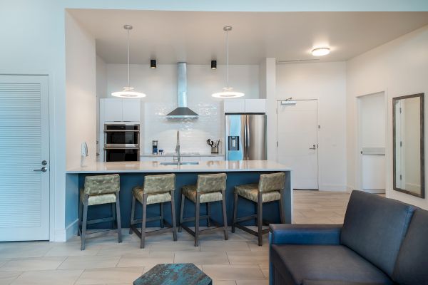 A modern kitchen with a central island, bar stools, stainless steel appliances, and pendant lighting. The space connects to a living area with a sofa.