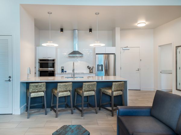 A modern kitchen with a central island, bar stools, stainless steel appliances, and pendant lighting. The space connects to a living area with a sofa.