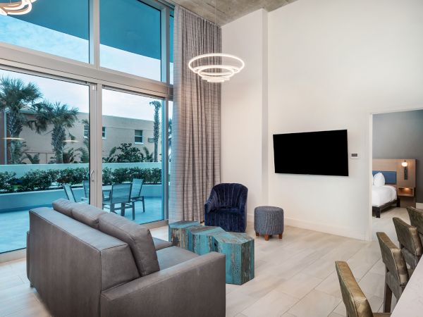 A modern living room with a couch, armchair, TV, and balcony access. There is a bedroom in the background. Large windows provide natural light.