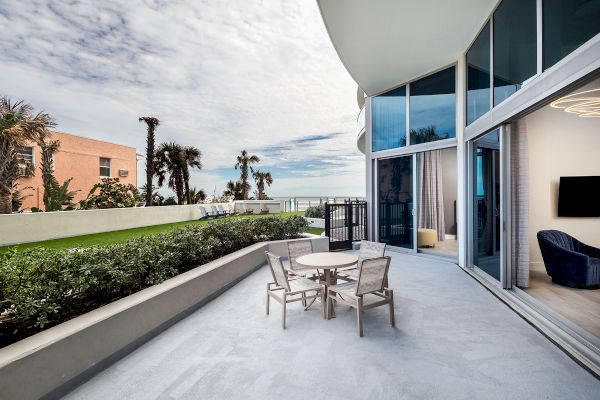 The image shows a modern balcony with a dining table and chairs overlooking a garden and ocean, adjacent to a glass-walled living area.