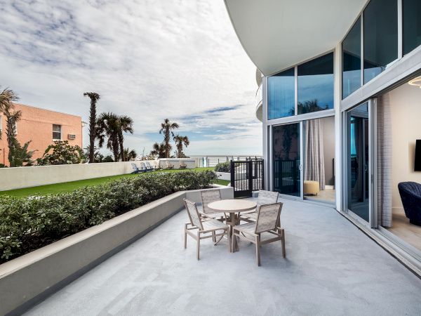 The image shows a modern balcony with a dining table and chairs overlooking a garden and ocean, adjacent to a glass-walled living area.